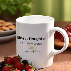 May include: White ceramic mug with the text "Eldest Daughter Family Manager Unpaid, Of Course". The mug is surrounded by fresh strawberries, blackberries, and blueberries. The mug has a curved handle.