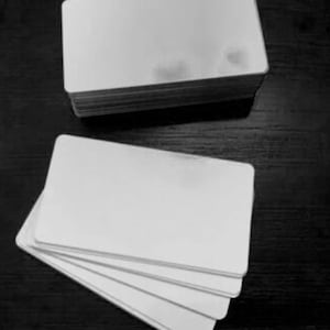 May include: A stack of blank, white rectangular cards, with rounded corners, sits atop a dark surface. Several additional cards are fanned out below the stack, showcasing the clean, smooth surface of the cards. The image is in black and white.