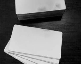 May include: A stack of blank, white rectangular cards, with rounded corners, sits atop a dark surface. Several additional cards are fanned out below the stack, showcasing the clean, smooth surface of the cards. The image is in black and white.