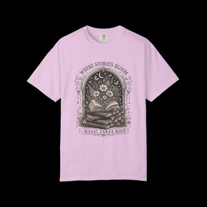 May include: A light pink short-sleeve t-shirt featuring a dark brown graphic of a stack of books with flowers blooming from the top book inside an arched frame. The text reads "WHERE STORIES BLOOM" at the top and "MAGIC TAKES ROOT" at the bottom.