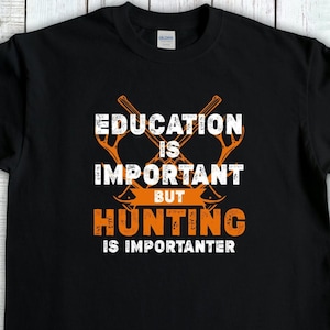 May include: Black t-shirt with the text "EDUCATION IS IMPORTANT BUT HUNTING IS IMPORTER". The design includes crossed hunting tools in orange and white lettering. The shirt is made of a soft material.