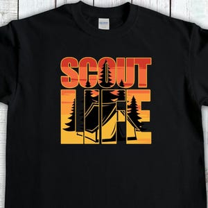 May include: Black t-shirt with the words "SCOUT LIFE" in orange and red block letters. The word "LIFE" features a camping scene with trees and a tent in a yellow and orange gradient.