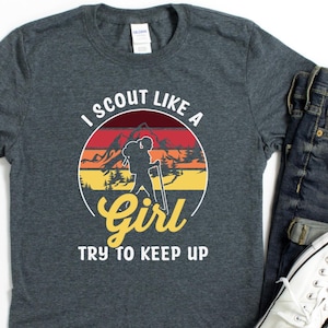 May include: Dark grey t-shirt with the text "I Scout Like A Girl Try To Keep Up" in a circular design featuring a hiker silhouette and mountain landscape. Accompanied by blue jeans and white trainers.