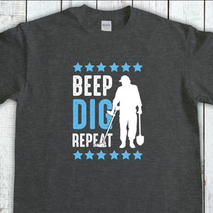 Op de afbeelding: Donkergrijs t-shirt met de tekst "BEEP DIG REPEAT" in wit en lichtblauw, samen met een silhouet van een persoon die met een metaaldetector zoekt. Blauwe sterren omringen de tekst. Het shirt is gemaakt van zacht materiaal.