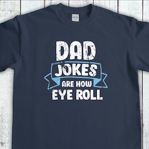 Peut inclure: T-shirt bleu marine avec l'inscription "DAD JOKES are how EYE ROLL" en lettres blanches et une bannière bleu clair. Le t-shirt est de style col rond.