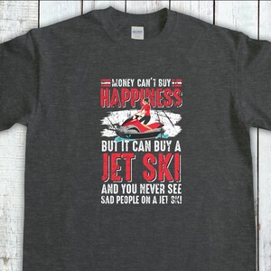 以下が含まれることがあります： 暗いグレーのTシャツに、白いグラフィックプリントが施されています。「お金で幸せは買えないけど、ジェットスキーは買えるし、ジェットスキーに乗ってる悲しい人を見たことがない！」と書いてあります。
