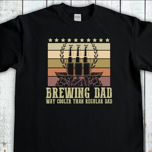 May include: Black t-shirt featuring a vintage-style graphic of beer bottles and hops. The text "Brewing Dad Way Cooler Than Regular Dad" is printed below the graphic.