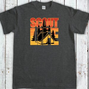 May include: Dark gray t-shirt with the words "SCOUT LIFE" in orange and yellow block letters. The design includes a black tent and trees, set against a sunset background. A Gildan tag is visible.