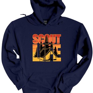 May include: Navy blue hooded sweatshirt with the words "SCOUT LIFE" in orange and yellow block letters. The design includes silhouettes of trees and a tent, evoking a camping theme. The sweatshirt has a front pocket and a drawstring hood.