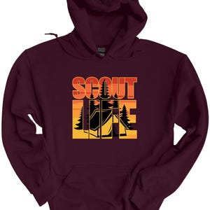 May include: A maroon hooded sweatshirt with the words "SCOUT LIFE" in orange and yellow block letters. The design includes a tent and trees, suggesting a camping or outdoor theme. The sweatshirt has a front pocket and a drawstring hood.
