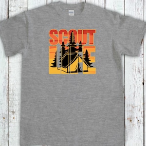 May include: A gray t-shirt featuring a camping-themed graphic. The design includes the word "SCOUT" in orange and red block letters above a black tent and trees set against a yellow and orange sunset.