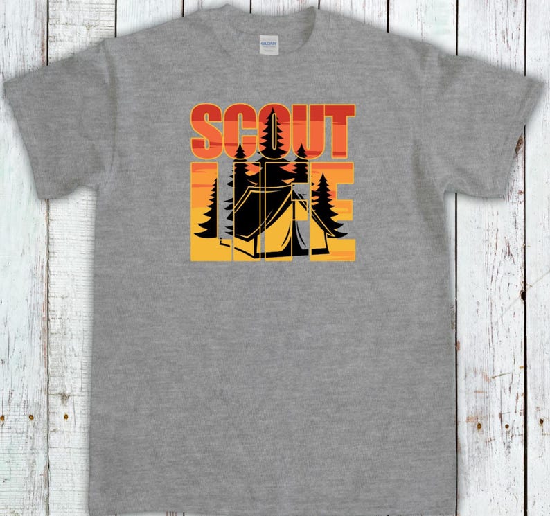May include: A gray t-shirt featuring a camping-themed graphic. The design includes the word "SCOUT" in orange and red block letters above a black tent and trees set against a yellow and orange sunset.