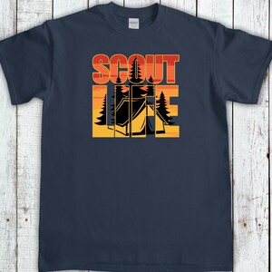 May include: Navy blue t-shirt with a camping-themed graphic. The design features the words "SCOUT LIFE" in orange and red block letters, with a silhouette of a tent and trees in the center. The shirt is made of a soft material.