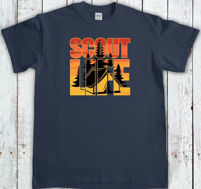 May include: Navy blue t-shirt with a camping-themed graphic. The design features the words "SCOUT LIFE" in orange and red block letters, with a silhouette of a tent and trees in the center. The shirt is made of a soft material.