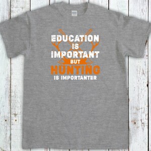 May include: A heather gray t-shirt with the text "EDUCATION IS IMPORTANT BUT HUNTING IS IMPORTER". The text is in white and orange, with crossed hunting tools above the word "HUNTING".