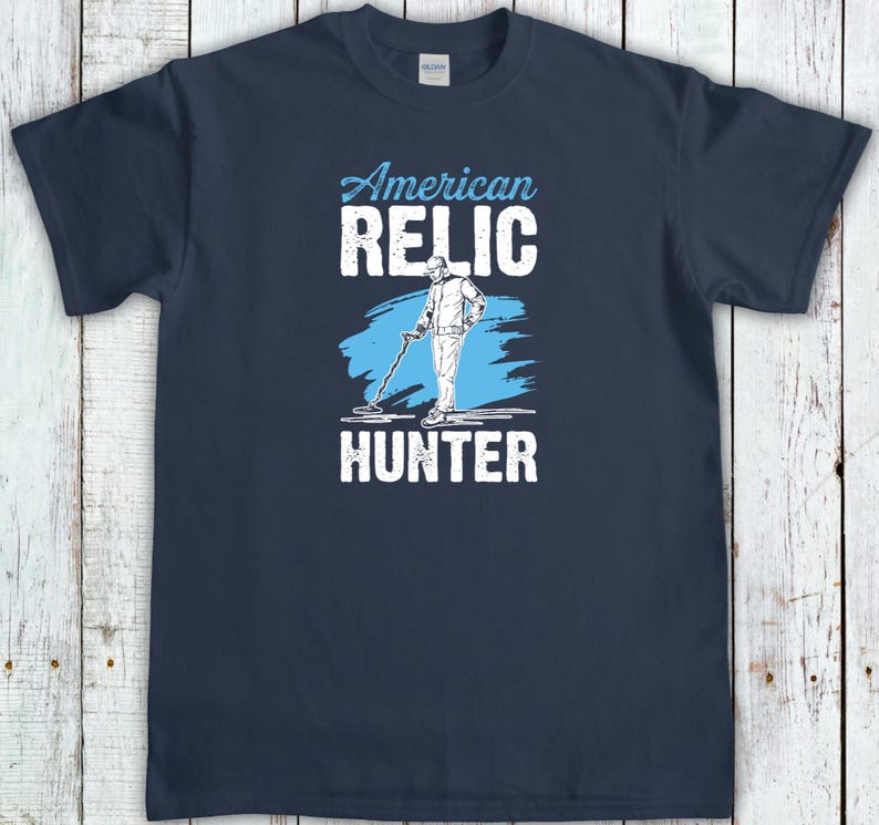 May include: Navy blue t-shirt with the words "American Relic Hunter" in white and light blue. The design features a graphic of a person using a metal detector. A great gift for treasure hunters.