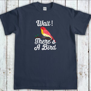 May include: Navy blue t-shirt with the text "Wait! There's A Bird" in white, featuring a colorful bird perched on a branch. The bird has red, yellow, and orange feathers. The shirt is made of a soft material.