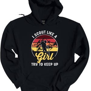 May include: Black hooded sweatshirt with a graphic design. The design features a hiker silhouette within a circular sunset scene, with the text "I SCOUT LIKE A Girl TRY TO KEEP UP" in white and yellow.