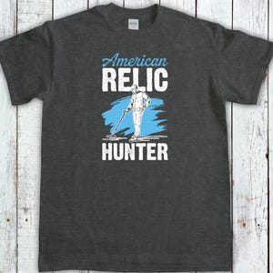 May include: Dark gray t-shirt with the words "American Relic Hunter" in white and light blue. The design features a person using a metal detector. The shirt is made of a soft, comfortable material.