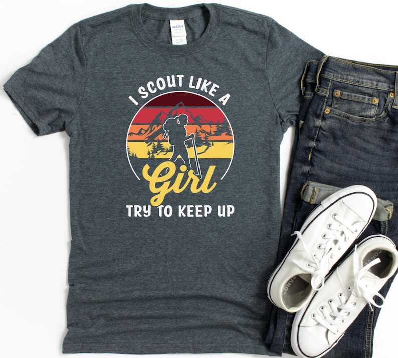 May include: Dark grey t-shirt with the text "I Scout Like A Girl Try To Keep Up" in a circular design featuring a hiker silhouette and mountain landscape. Accompanied by blue jeans and white trainers.