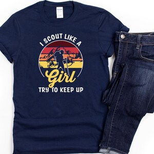 May include: Navy blue t-shirt with a circular graphic featuring a hiker silhouette, mountains, and the text "I SCOUT LIKE A GIRL TRY TO KEEP UP." The shirt is paired with dark wash denim jeans.