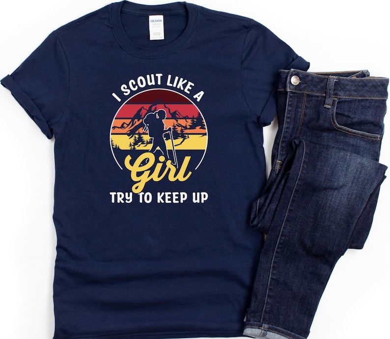 May include: Navy blue t-shirt with a circular graphic featuring a hiker silhouette, mountains, and the text "I SCOUT LIKE A GIRL TRY TO KEEP UP." The shirt is paired with dark wash denim jeans.