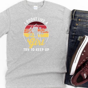 May include: A grey t-shirt featuring the text "I Scout Like a Girl, Try to Keep Up" in a circular design with a mountain and sunset graphic. The shirt is shown with blue jeans and maroon high-top trainers.