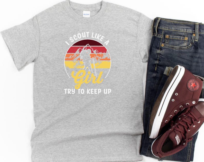 May include: A grey t-shirt featuring the text "I Scout Like a Girl, Try to Keep Up" in a circular design with a mountain and sunset graphic. The shirt is shown with blue jeans and maroon high-top trainers.