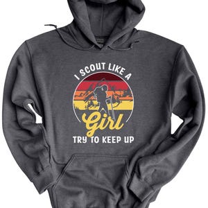 May include: Dark grey hooded sweatshirt with a front pocket and drawstring. The front features a circular graphic with a hiker silhouette, mountains, and the text "I SCOUT LIKE A Girl TRY TO KEEP UP."
