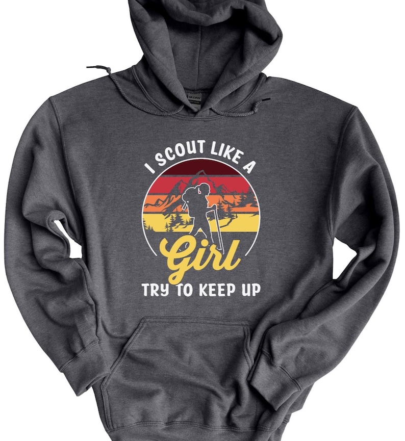 May include: Dark grey hooded sweatshirt with a front pocket and drawstring. The front features a circular graphic with a hiker silhouette, mountains, and the text "I SCOUT LIKE A Girl TRY TO KEEP UP."