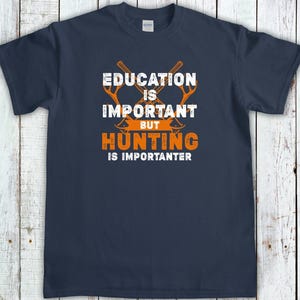May include: Navy blue t-shirt with the text "EDUCATION IS IMPORTANT BUT HUNTING IS IMPORANTER" in white and orange. The design includes two crossed axes. The shirt is made of a soft material.