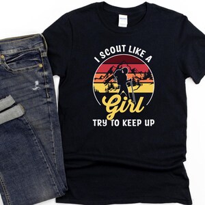 May include: Black t-shirt with the text "I Scout Like a Girl Try to Keep Up" in white and yellow. The design features a silhouette of a person hiking with mountains and a sunset background. A pair of blue jeans is next to the shirt.