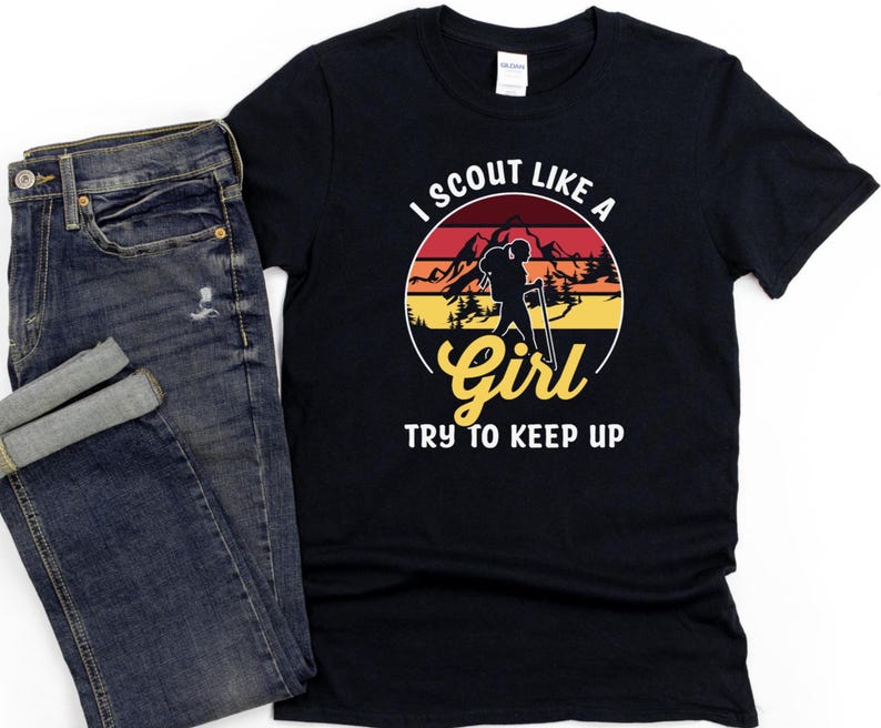May include: Black t-shirt with the text "I Scout Like a Girl Try to Keep Up" in white and yellow. The design features a silhouette of a person hiking with mountains and a sunset background. A pair of blue jeans is next to the shirt.