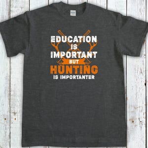 May include: Dark gray t-shirt with the text "EDUCATION IS IMPORTANT BUT HUNTING IS IMPORANTER" in white and orange. The design includes crossed axes. The shirt is made of a soft material.