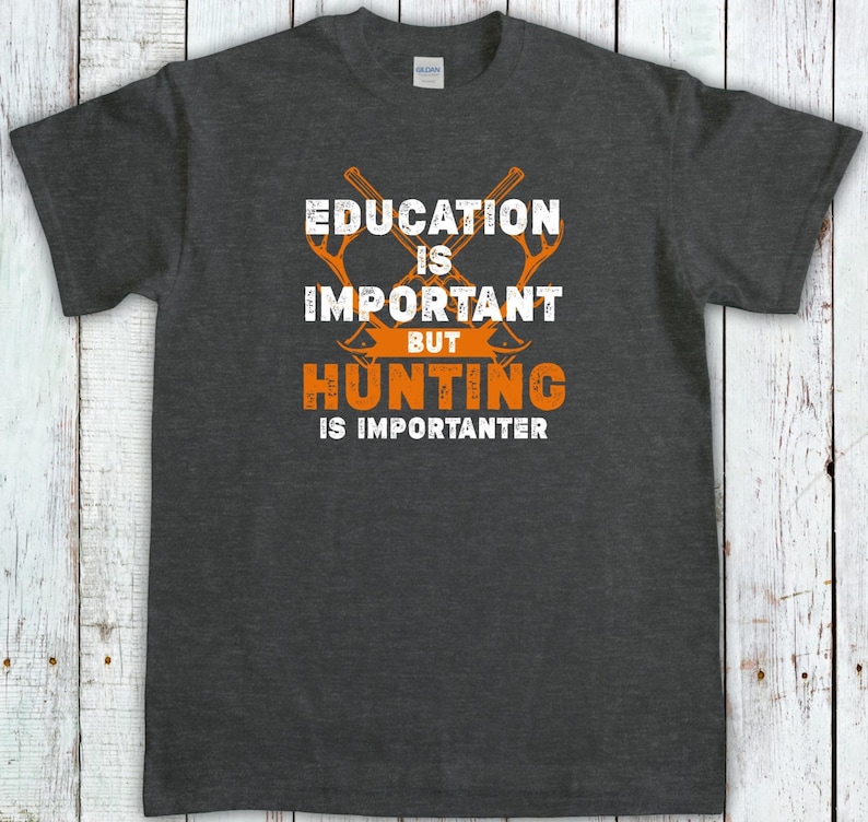 May include: Dark gray t-shirt with the text "EDUCATION IS IMPORTANT BUT HUNTING IS IMPORANTER" in white and orange. The design includes crossed axes. The shirt is made of a soft material.