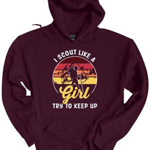 May include: A maroon hoodie with a graphic design. The design features a silhouette of a person hiking with a backpack, set against a sunset backdrop with the text "I SCOUT LIKE A GIRL TRY TO KEEP UP."