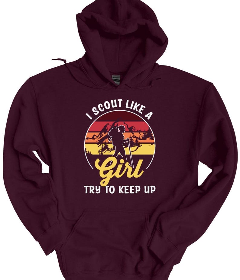 May include: A maroon hoodie with a graphic design. The design features a silhouette of a person hiking with a backpack, set against a sunset backdrop with the text "I SCOUT LIKE A GIRL TRY TO KEEP UP."