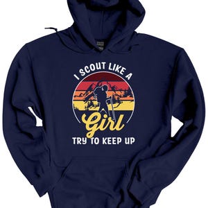 May include: A navy blue hoodie with a graphic that reads "I Scout Like a Girl, Try to Keep Up." The graphic features a silhouette of a hiker with a backpack and walking stick in front of a mountain range and a sunset.