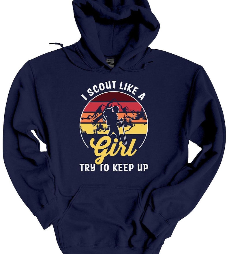 May include: A navy blue hoodie with a graphic that reads "I Scout Like a Girl, Try to Keep Up." The graphic features a silhouette of a hiker with a backpack and walking stick in front of a mountain range and a sunset.