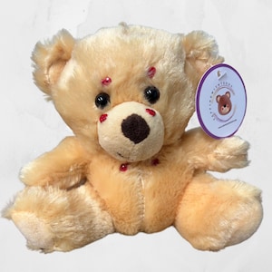 May include: A small, beige plush teddy bear with red felt accents on its face and body. The bear is sitting with its legs crossed and has a small tag attached to its right ear that says "Little Adventures".