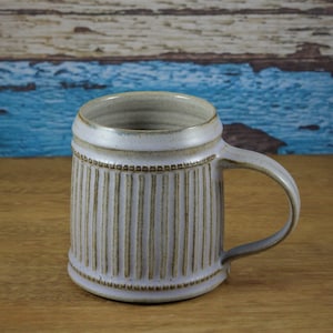 May include: A light grey ceramic mug with a ribbed design and a handle. The mug has a subtle brown glaze.