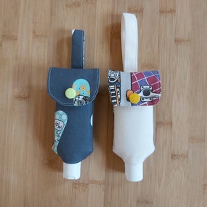 Könnte beinhalten: Zwei Stoff-Handdesinfektionsmittel-Halter mit einer Schlaufe zum Aufhängen. Ein Halter ist grau mit weißen Punkten und einem blauen und grünen Druck. Der andere Halter ist weiß mit einem bunten Druck von Skateboards. Beide Halter haben einen gelben Knopfverschluss.
