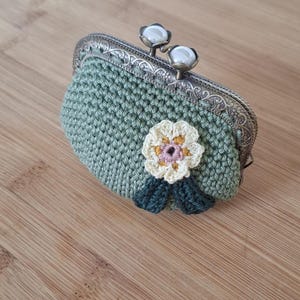 Puede incluir: Un monedero de ganchillo en un tono verde suave, adornado con una flor color crema con detalles en rosa y amarillo y hojas verdes. El monedero presenta un cierre de metal plateado.