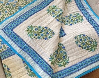 May include: A quilted throw blanket featuring an off-white base with a repeating green and blue floral block print pattern. The blanket is finished with a bright blue border and a secondary blue and white chevron-style trim along the edges.