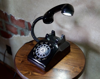 May include: A black rotary phone with a built-in lamp. The phone has a black dial with white numbers and a white bell icon. The lamp is attached to the phone's handset and has a white light bulb.