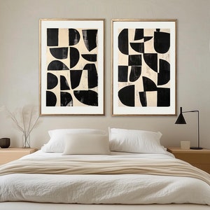 May include: Two framed abstract art prints with black and white geometric shapes on a cream background. The prints are hanging above a white bed with a beige throw blanket.