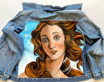 May include: A denim jacket with a painting of the Birth of Venus on the back. The painting is done in a realistic style and features a woman with long flowing hair and a soft, ethereal expression.