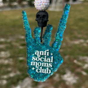 May include: A hanging ornament shaped like a hand making a rock-and-roll gesture, crafted from teal glitter resin. It features the white text "anti social moms club" with small star accents, topped with a black skull bead and a white iridescent bead.