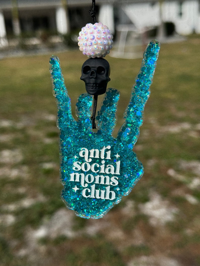 May include: A hanging ornament shaped like a hand making a rock-and-roll gesture, crafted from teal glitter resin. It features the white text "anti social moms club" with small star accents, topped with a black skull bead and a white iridescent bead.