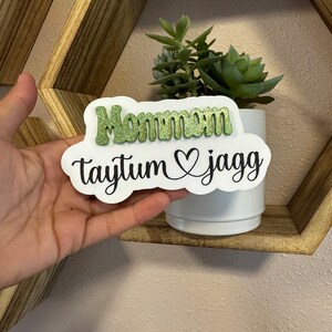 May include: A white sign with the words "Mommom Taytum & Jagg" in black lettering. The word "Mommom" is in green glitter.